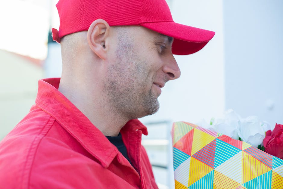 A man dressed in a red uniform and matching red cap holding a floral bouquet wrapped in colorful geometric-patterned paper, featuring white and red roses, with a soft natural light illuminating the sc