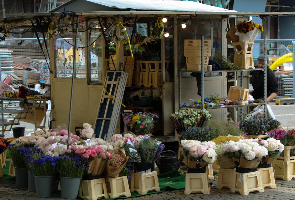 A bustling outdoor flower stall featuring an array of fresh floral arrangements. Numerous buckets and bouquets of pink, white, purple, and blue flowers are displayed on beige wooden crates and black c