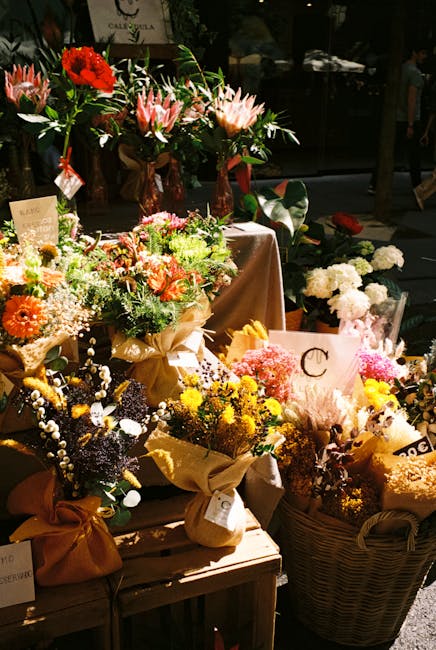 A diverse display of fresh flower arrangements and bouquets arranged on a wooden table and baskets. The flowers include pink lilies, bright red roses, white and green chrysanthemums, and small pastel-