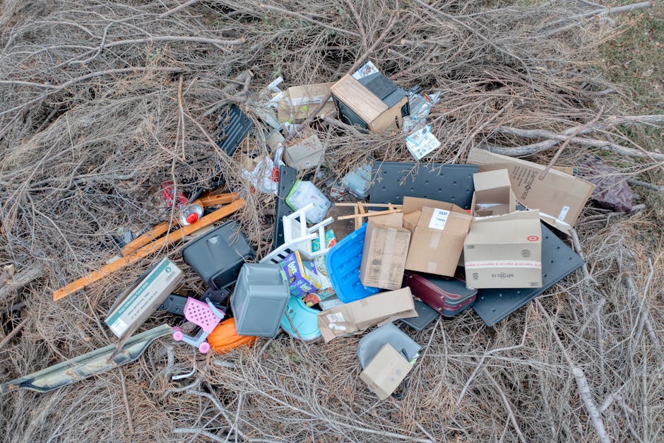 An aerial view of a pile of miscellaneous household waste including cardboard boxes, plastic containers, a woven basket, and miscellaneous packaging materials, scattered amidst dry branches and twigs 