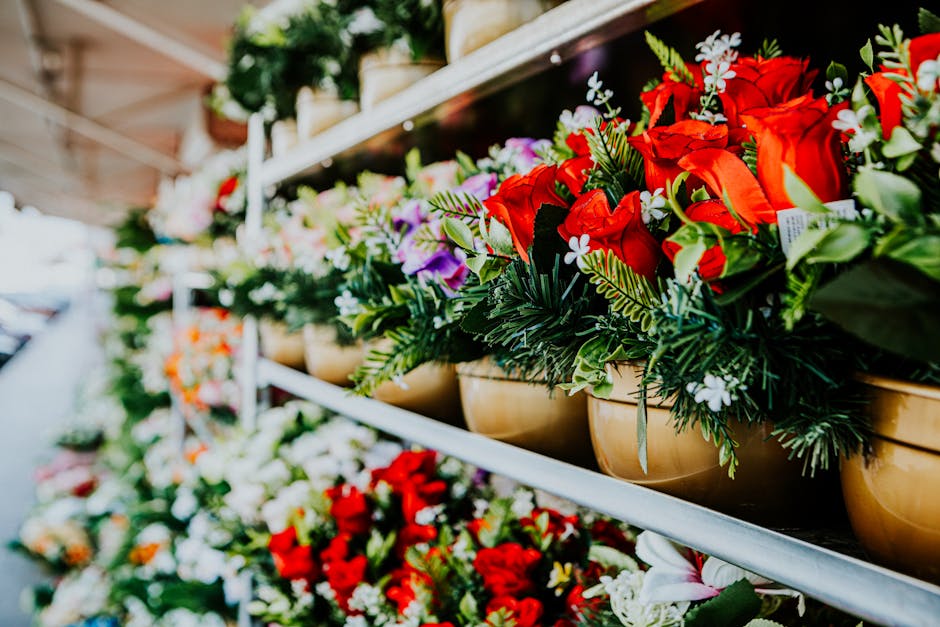A close-up view of a floral arrangement in a beige ceramic pot featuring vibrant red roses, white flowers, and green foliage, displayed on a metal shelf at a flower market. The bouquet showcases fresh
