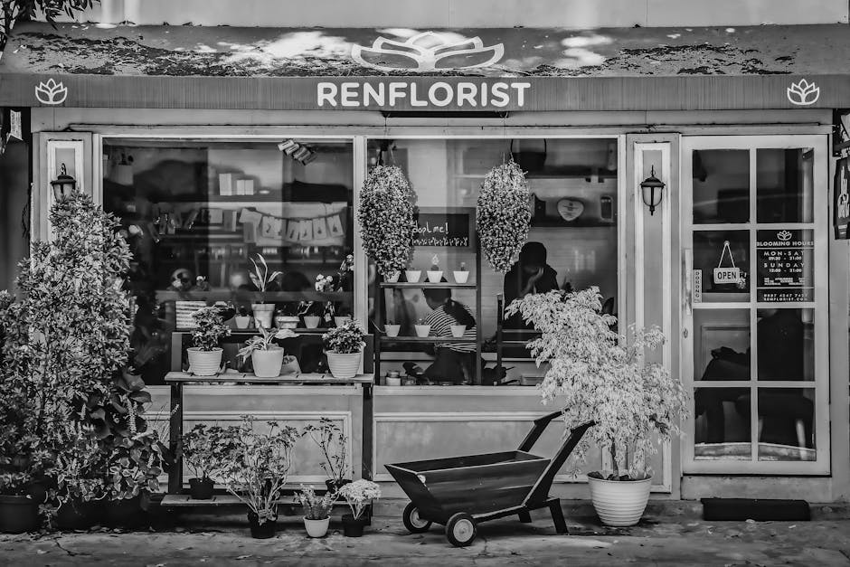Black and white photograph of the exterior of Renflorist, a flower shop with a wooden signboard displaying their name. The shop's window showcases an arrangement of potted plants and hanging floral de
