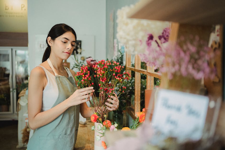 A young woman with dark hair tied back, wearing a white sleeveless top and a beige apron, arranging a bouquet of small pink and red hypericum berries and flowers in a floral shop. She is holding the b