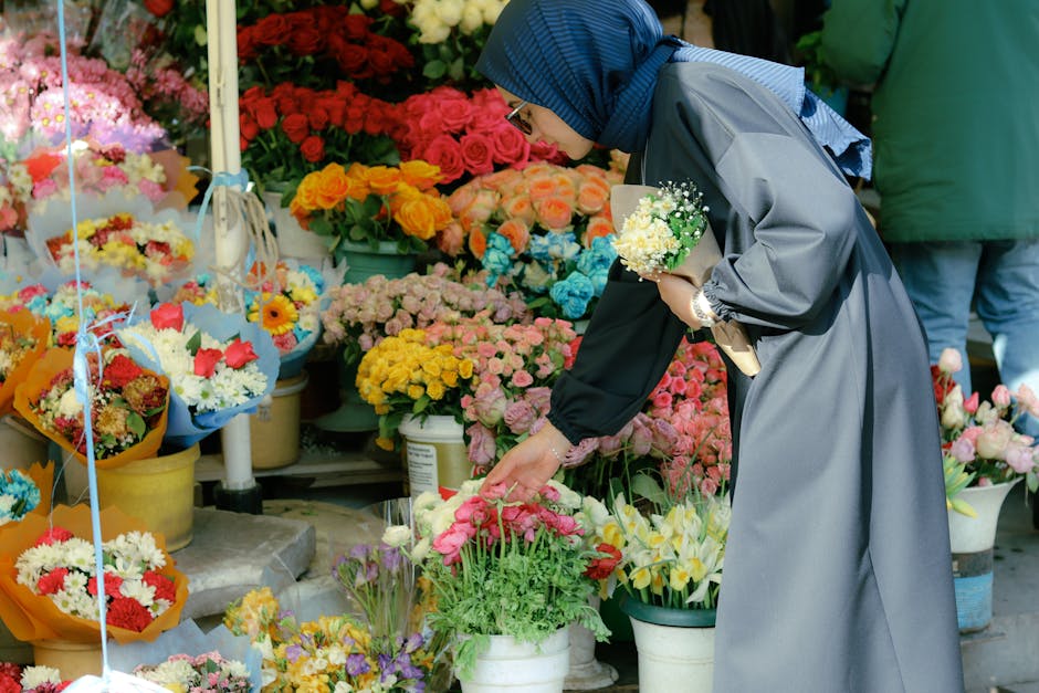 A woman wearing a blue hijab, glasses, and a long grey coat is shopping for fresh flowers at a bustling outdoor flower market. She is leaning forward, carefully selecting a vibrant bouquet of pink and