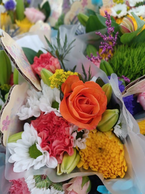 A close-up of a floral bouquet arranged with a variety of fresh flowers, including an orange rose as the central focal point, surrounded by white daisies with green centers, pink carnations, yellow ch