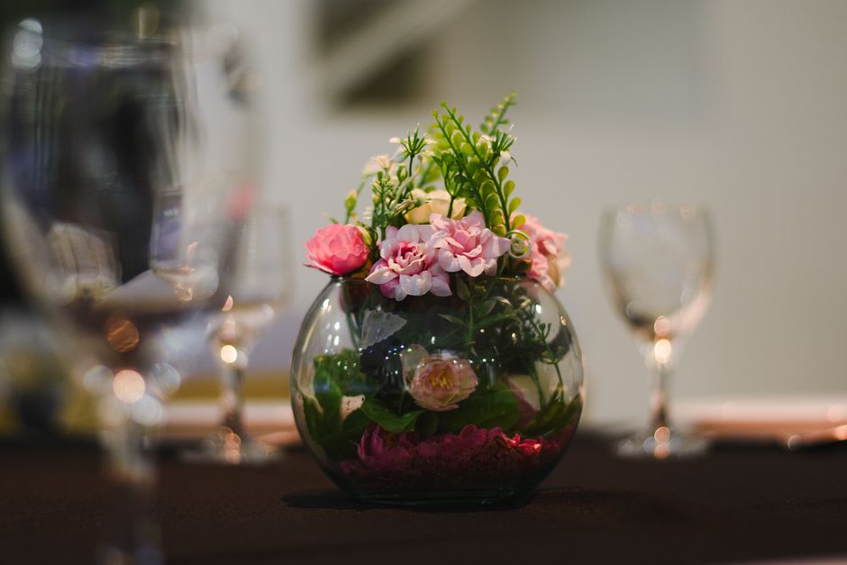 A round glass vase filled with a floral arrangement of pink and white flowers, including roses and hydrangeas, with green foliage and succulent-like stems. The flowers are fresh, with delicate petals 