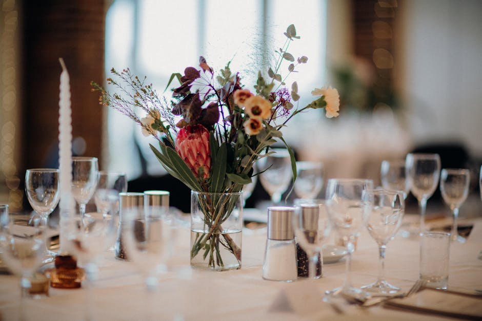 A floral arrangement featuring a variety of fresh flowers including a prominent pink protea, small cream and purple daisies, purple eucalyptus leaves, and delicate purple filler flowers, all arranged 
