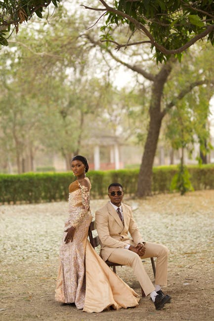 A bride and groom pose in a park setting beneath the leafy branches of a large tree, with lush green grass and trimmed hedges in the background. The bride wears an elegant off-the-shoulder lace gown i