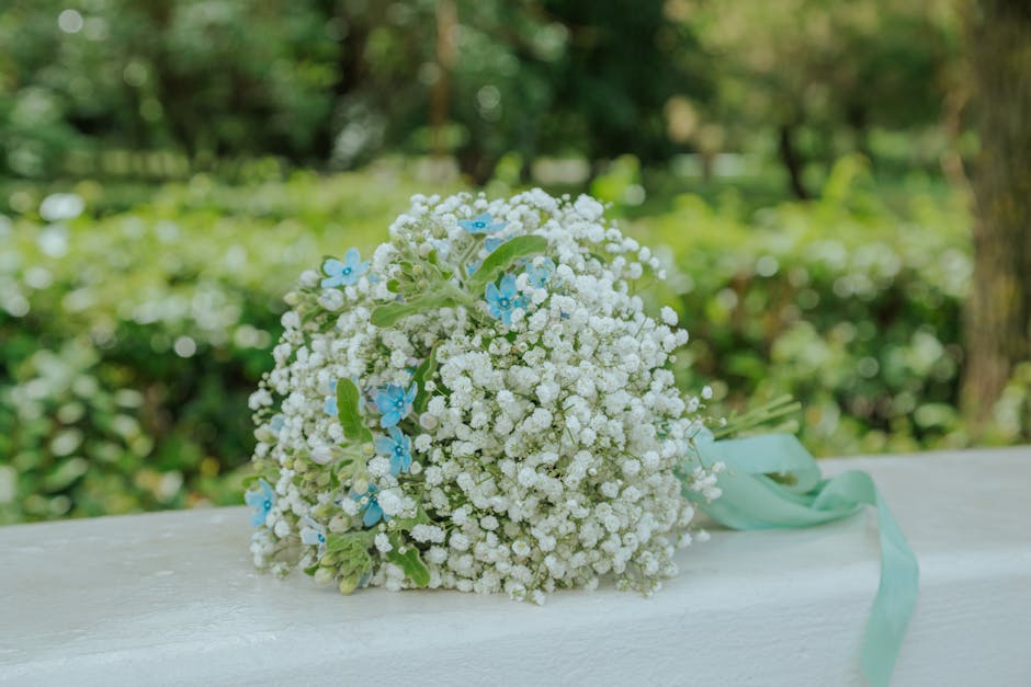 A bouquet of fresh, delicate baby's breath flowers, accented with small blue forget-me-not blooms and light green foliage, tied together with a soft aqua ribbon. The bouquet rests on a white wooden su