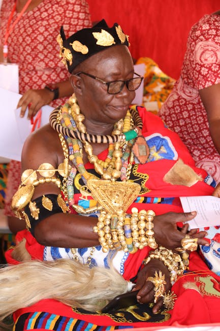 A woman dressed in traditional African attire with vibrant red and multicolored woven fabrics, adorned with elaborate gold jewelry including necklaces, bracelets, and earrings. She is wearing a black