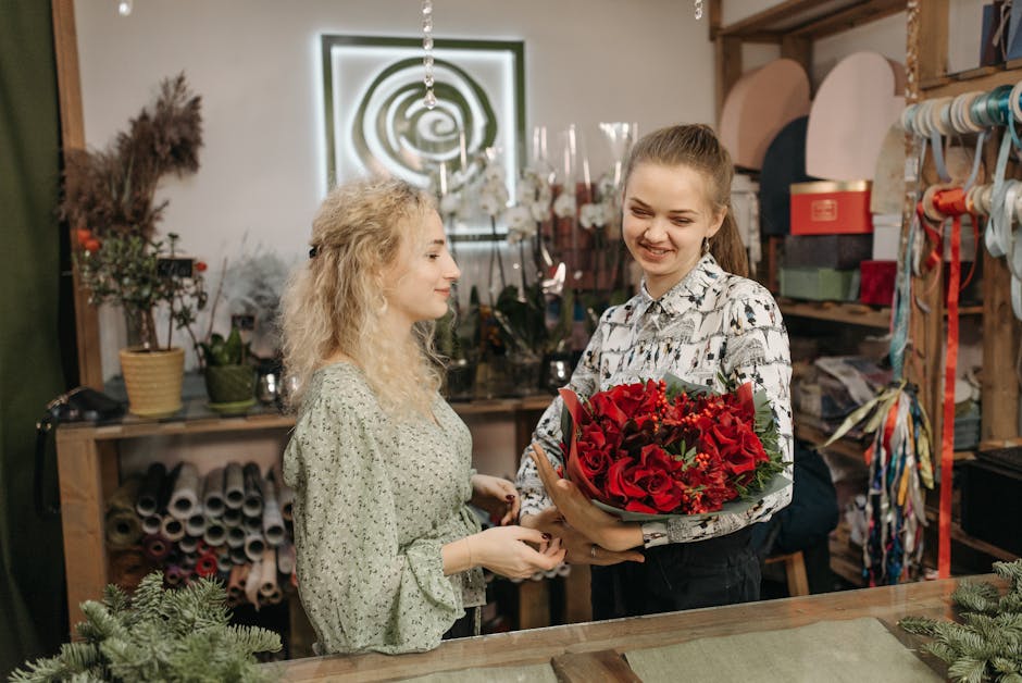 A florist in a flower shop holding a vibrant bouquet of red roses, lilies, and greenery wrapped in brown paper and bordered with dark green leaves. She is smiling as she accepts the floral gift from a